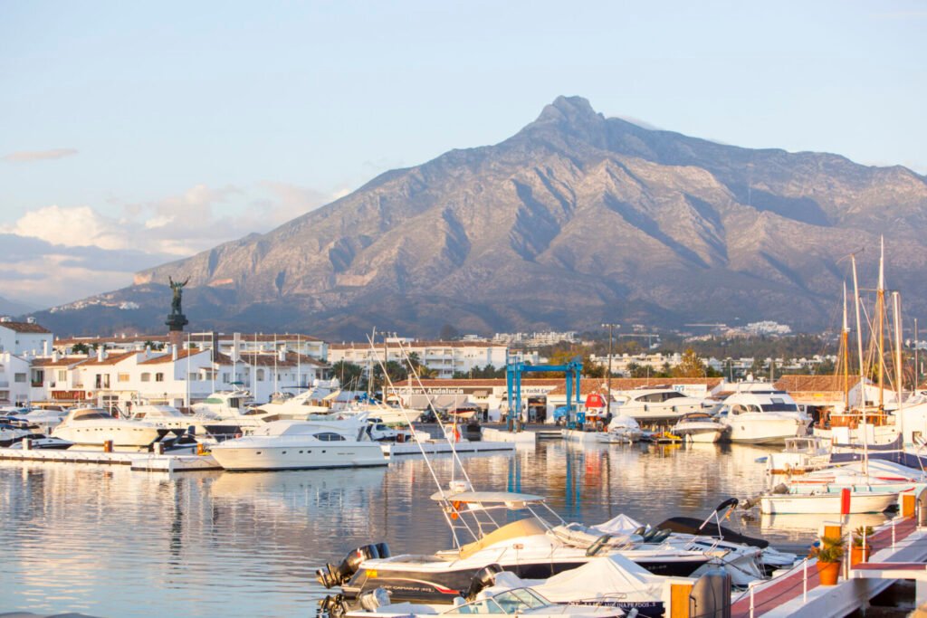 Luxury yachts docked at Puerto Banús marina in Marbella with La Concha mountain in the background on a sunny day.
