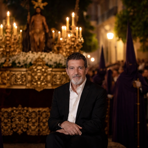 Antonio Banderas depicted during a Semana Santa procession with a candlelit float and nazarenos in purple robes in the background.