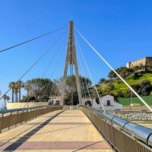Modern suspension bridge in Fuengirola leading towards the historic Sohail Castle on a sunny day.