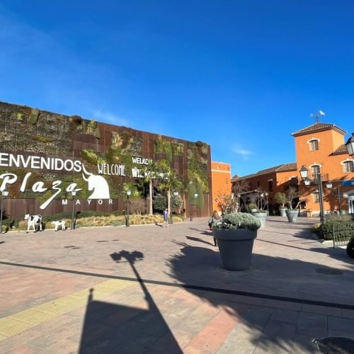 Outdoor view of Plaza Mayor Málaga shopping centre with welcome sign, open square, palm trees and traditional Andalusian-style buildings under a blue sky.